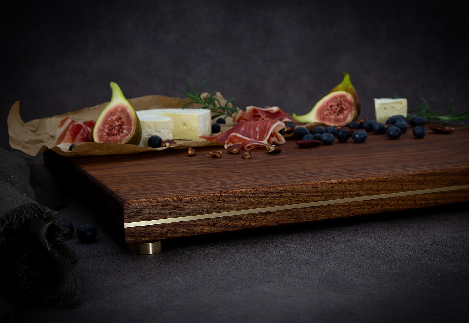 A HEWN chopping and serving board in Tasmanian Blackwood, brass inlaid along its edge and raised on brass feet, with figs, brie, prosciutto and blueberries