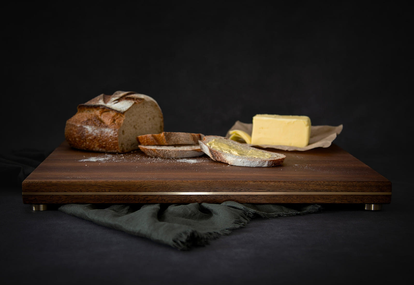 A HEWN chopping and serving board in Spotted Gum, brass inlaid along its edge and raised on brass feet, holding a sourdough loaf, sliced bread and butter